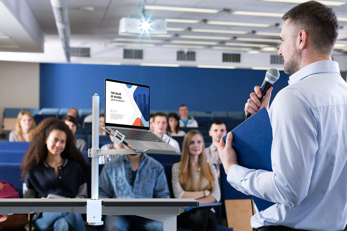 Lecturer presenting to a hall with a laptop on a pole-clamp mount at the desk
