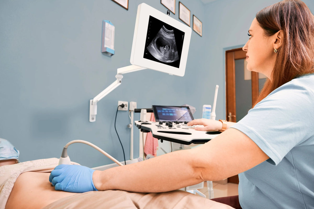 Sonographer with a display on an articulating VESA arm beside the exam table